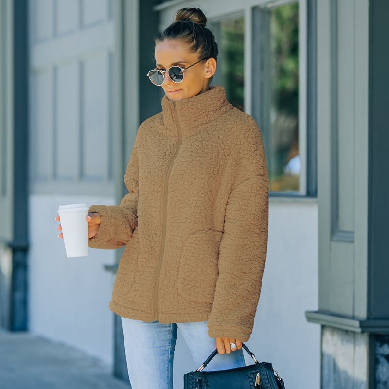 Frau in brauner Fleecejacke mit Kaffeebecher und Handtasche, draußen stehend.