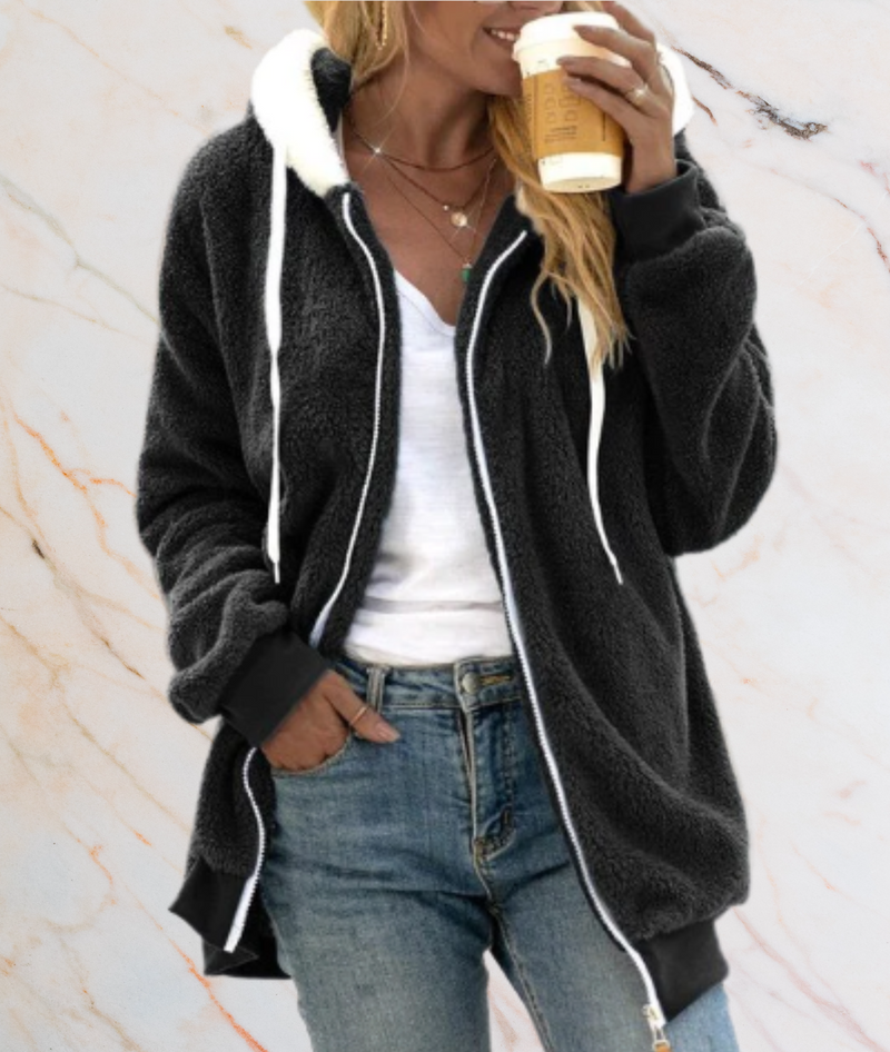 Frau in schwarzer Fleecejacke mit Kapuze, hält Kaffeebecher, lässiger Herbstlook.