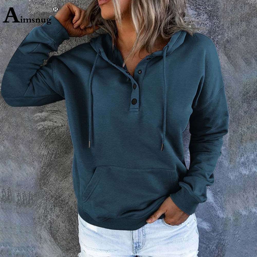 Frau in dunkelblauem Kapuzenpullover mit Knöpfen und Kängurutasche vor grauer Wand.