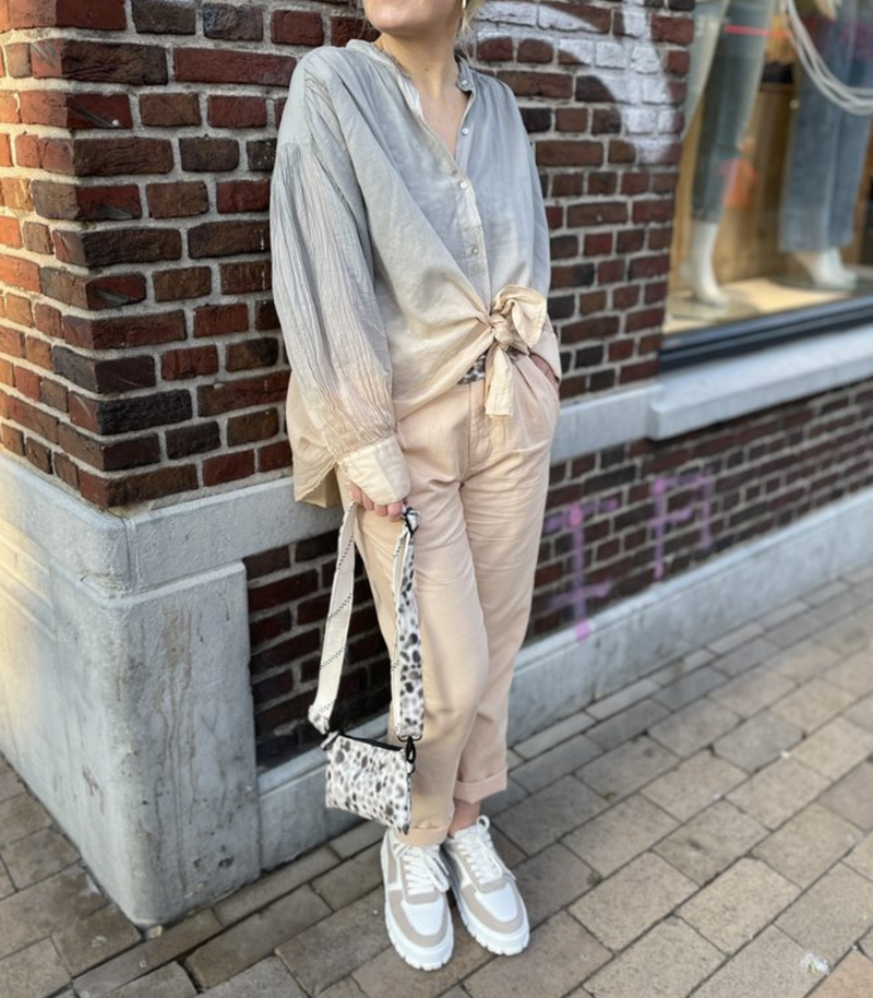 Frau in beiger Hose und grauer Bluse mit Leoparden-Tasche, steht vor Backsteinwand.