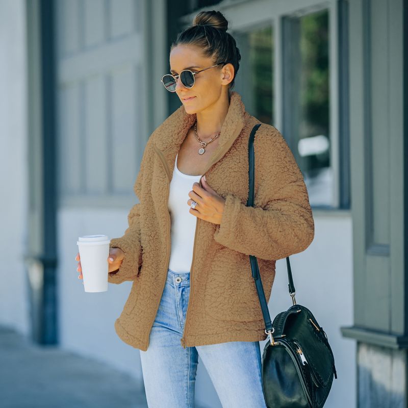 Frau in brauner Teddyjacke, Sonnenbrille, Jeans, hält Kaffeebecher, modisch, urbaner Stil.