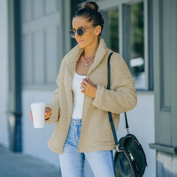 Frau in beiger Teddyjacke, Sonnenbrille, Jeans, mit Kaffeebecher und schwarzer Tasche.