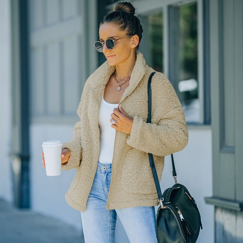 Frau in beiger Teddyjacke, Sonnenbrille, Jeans, mit Kaffeebecher und schwarzer Tasche.