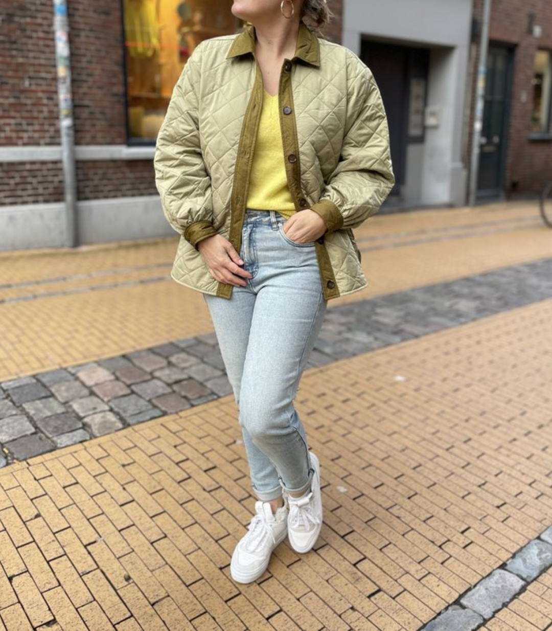 Frau in olivgrüner Steppjacke, gelbem Pullover, blauer Jeans und weißen Sneakers auf Straße.