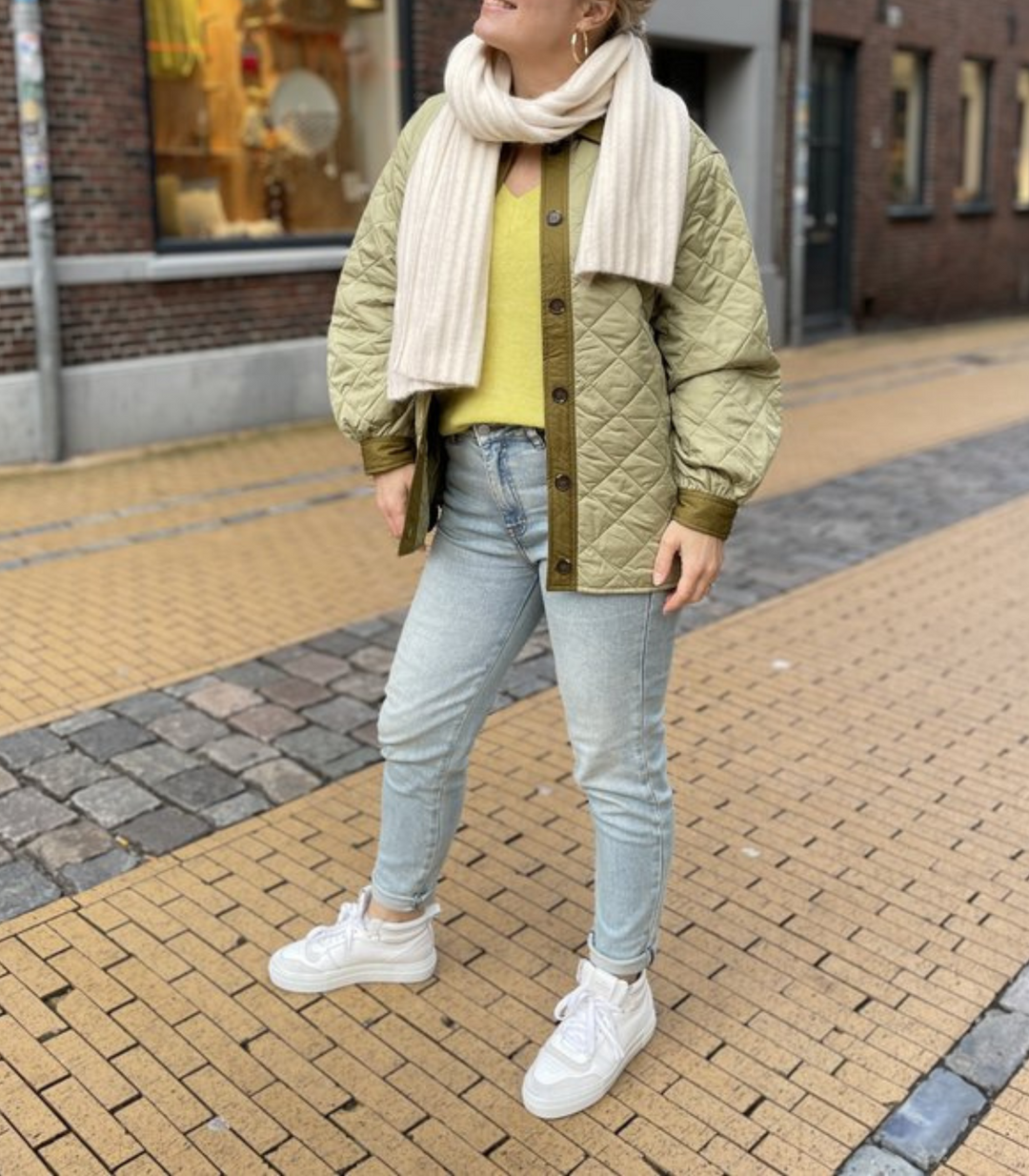 Frau in olivgrüner Steppjacke, gelbem Pullover, blauer Jeans und weißen Sneakers auf Straße.