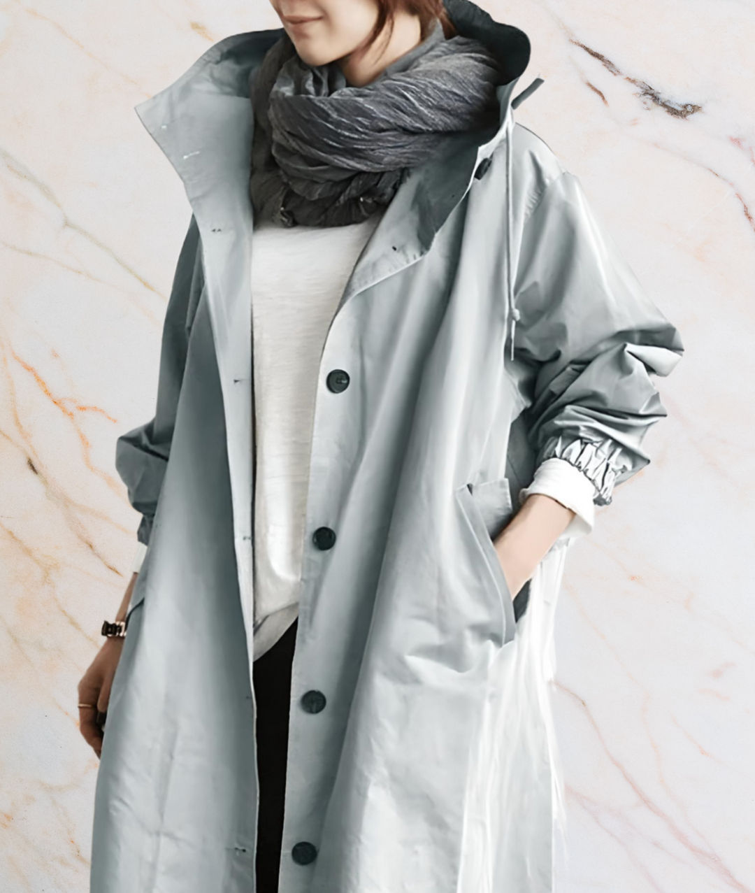 Frau in grauem Regenmantel mit Schal vor Marmorhintergrund.