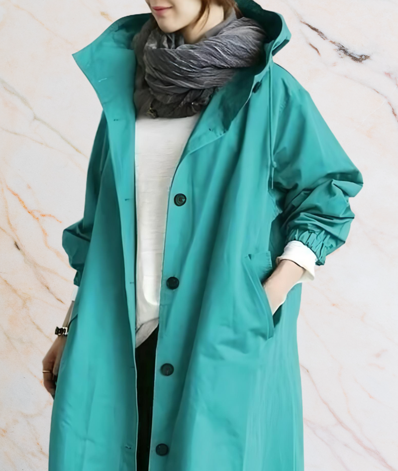 Frau in türkisfarbenem Regenmantel mit Kapuze und grauem Schal vor Marmorhintergrund.