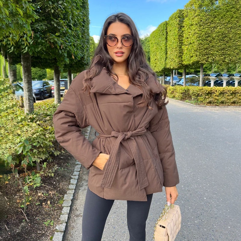 Frau in brauner Daunenjacke mit Gürtel, Sonnenbrille, schwarze Hose, beige Handtasche, draußen.