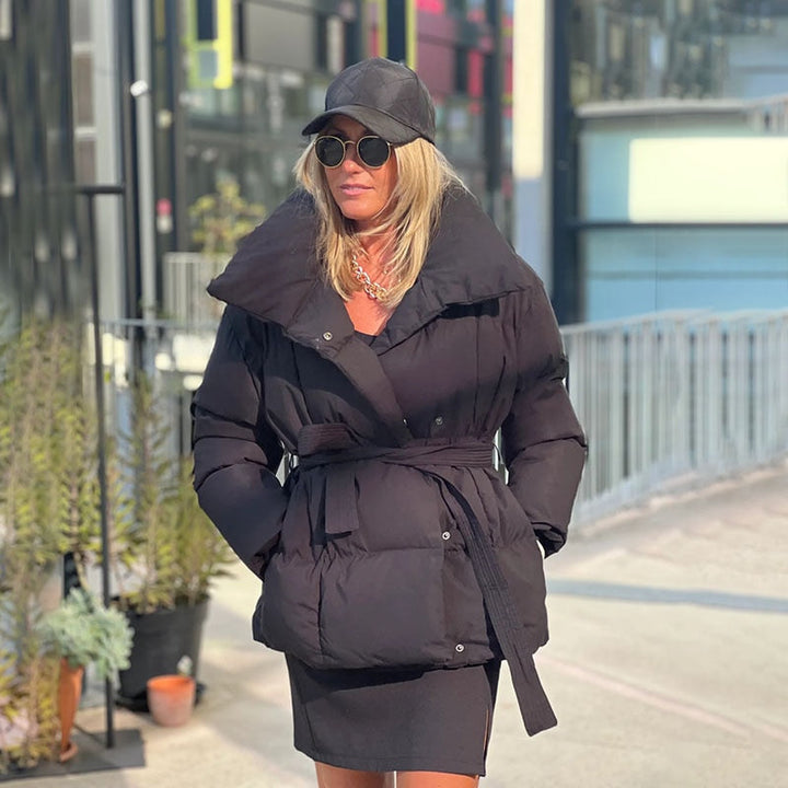 Frau in schwarzer Daunenjacke, Sonnenbrille und Mütze, modisch, urbaner Hintergrund.