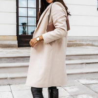 Frau in beigem Wollmantel, schwarze Lederhose, braune Tasche, Armbanduhr, draußen.