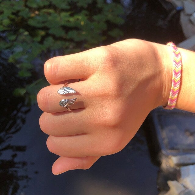 Hand mit silbernem Engelsflügel-Ring und bunter Freundschaftsarmband im Sonnenlicht.