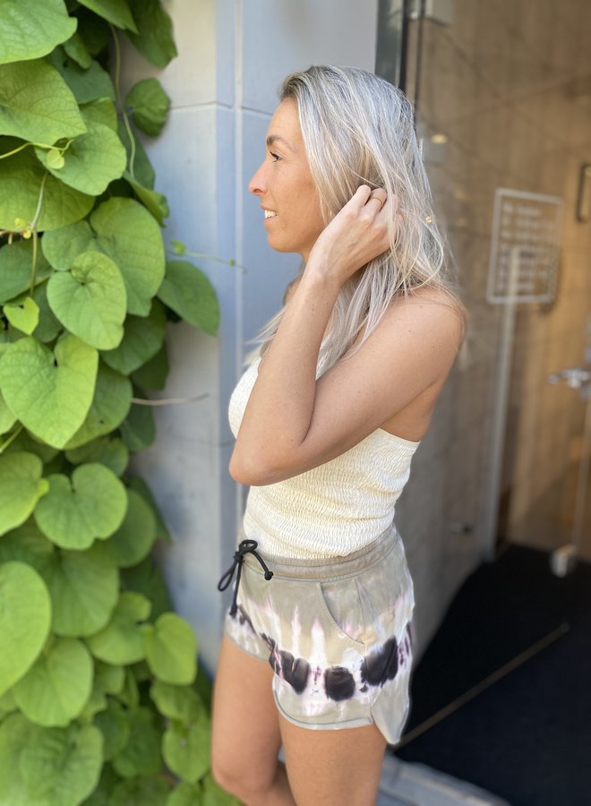 Frau in cremefarbenem Top und gemusterten Shorts, stehend neben grünen Blättern.