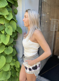 Frau in weißem Tanktop und gemusterten Shorts neben grüner Pflanze, lächelnd, seitlich.