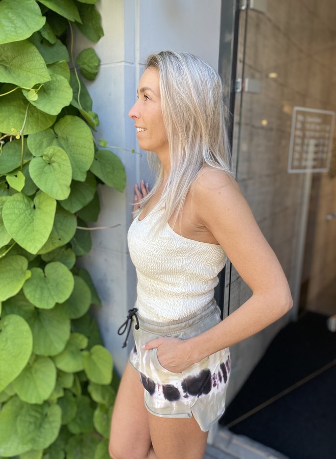 Frau in weißem Tanktop und gemusterten Shorts neben grüner Pflanze, lächelnd, seitlich.