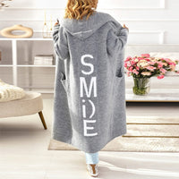 Frau in grauem Kapuzen-Cardigan mit "SMILE"-Aufdruck, Wohnzimmer, Blumenstrauß, gemütlich.