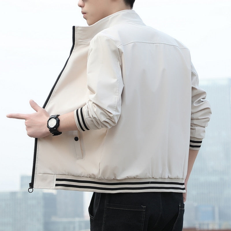 Mann in beiger Bomberjacke mit schwarzen Streifen, Uhr am Handgelenk, urbaner Hintergrund.