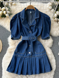 Blaues Jeanskleid mit Gürtel, Puffärmeln und Faltenrock auf weißem Fellteppich.