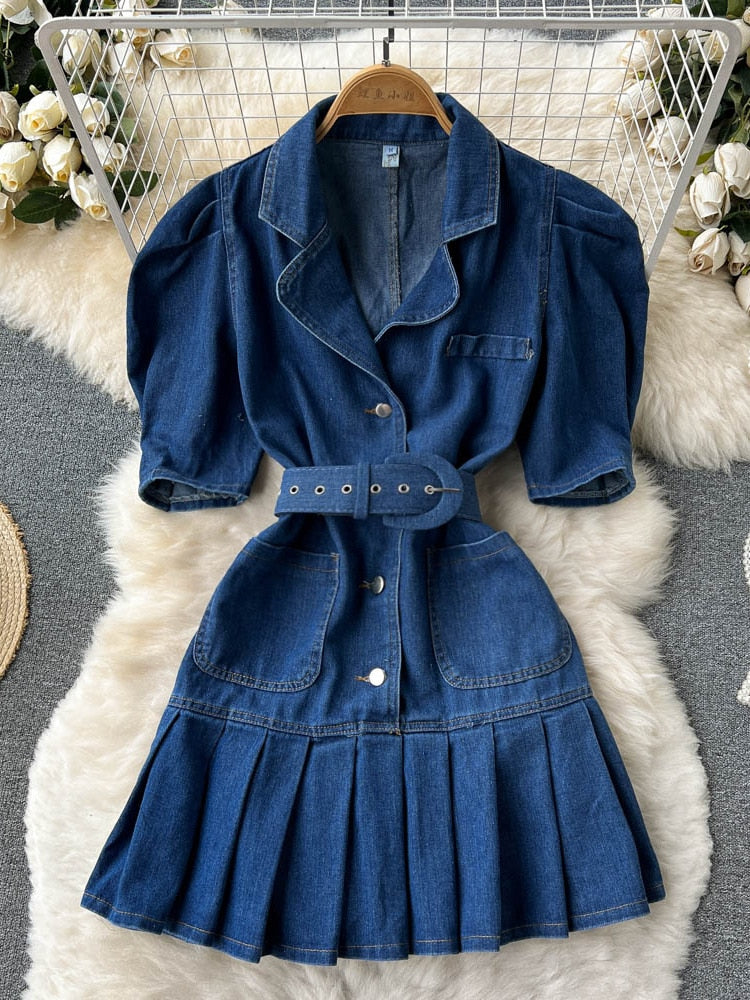 Blaues Jeanskleid mit Gürtel, Puffärmeln und Faltenrock auf weißem Fellteppich.