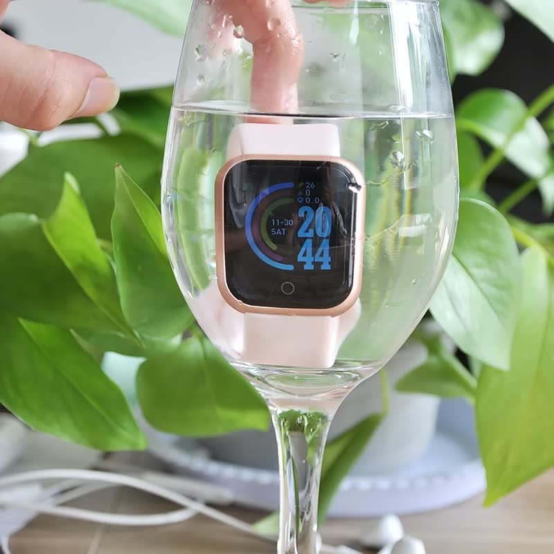 Wasserdichte Smartwatch in Roségold, im Weinglas, mit digitalem Display, vor grünen Blättern.