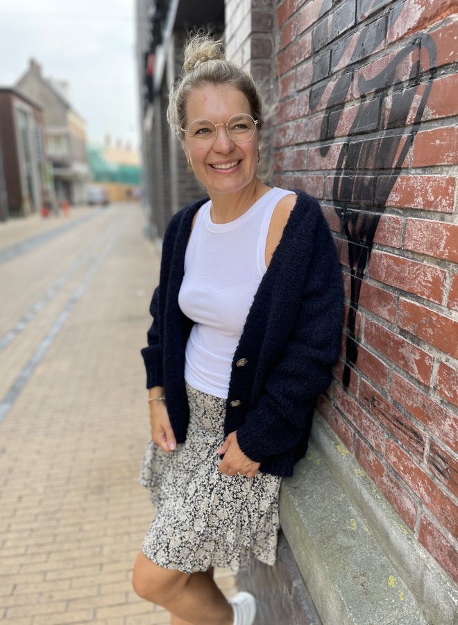 Frau in weißem Top, schwarzer Strickjacke und gemustertem Rock lehnt an Ziegelwand.