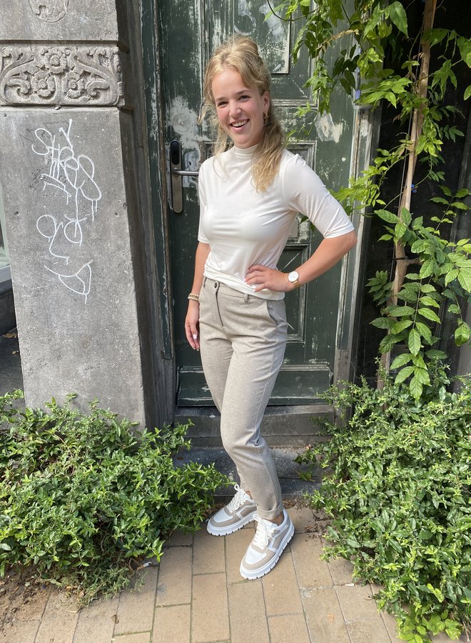 Frau in weißem Rollkragen-T-Shirt und grauer Hose vor grüner Tür, lächelnd, draußen.