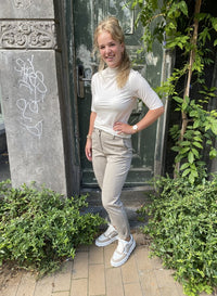 Frau in weißem Rollkragen-T-Shirt und grauer Hose vor grüner Tür, lächelnd, draußen.